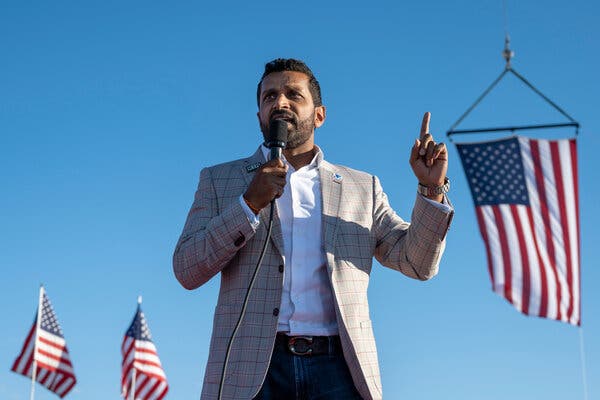 Kash Patel speaking outdoors into a microphone in front of three American flags.