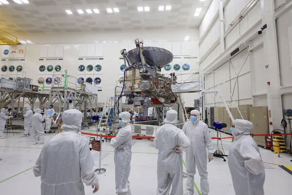 Several people in white jumpsuits and facemasks stand around in a large bright clean room with the spacecraft looming in the background.