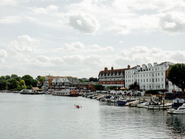 Two people kayak in a wide expanse of river beside boats that are docked in front of buildings and a cloudy sky.