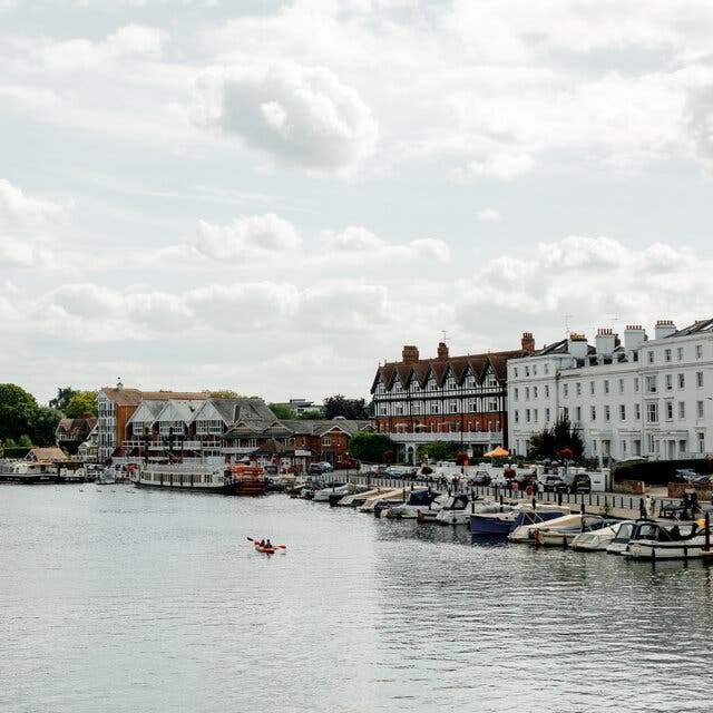 Two people kayak in a wide expanse of river beside boats that are docked in front of buildings and a cloudy sky.