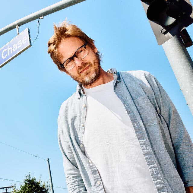 Shot from below, a man with tussled hair, a beard and glasses looks downward. Behind him is a bright blue sky, the long metal arm of a traffic light and a street sign that reads “Chevy Chase.”