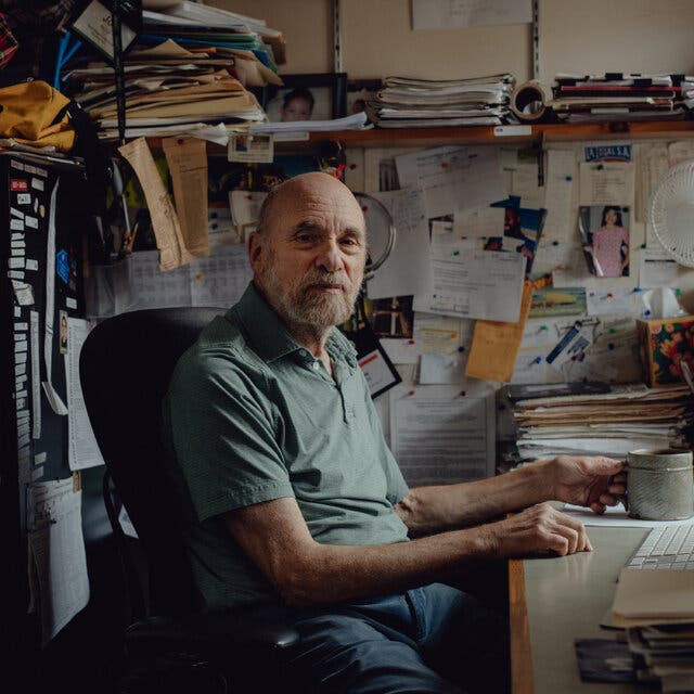 A bald man in a green polo shirt sits at a desk with a bulletin board crammed with notices behind him.