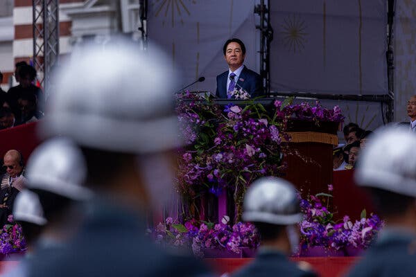 A man in a suit speaks at a podium bedecked with purple flowers. In the foreground, out of focus, are soldiers in helmets.