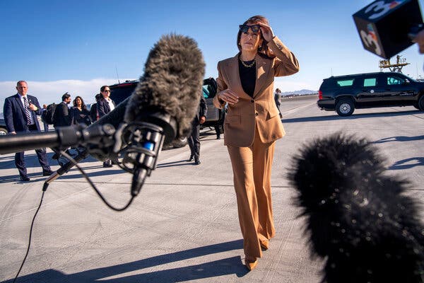 Kamala Harris raises her sunglasses while walking toward a bank of microphones set up on an airport tarmac.