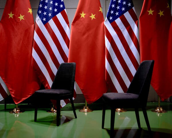 Two empty chairs in room, with a row of U.S. and Chinese flags behind them.