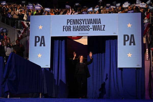 Kamala Harris enters a stage from underneath a sign that says Pennsylvania for Harris-Walz with a crowd behind her.