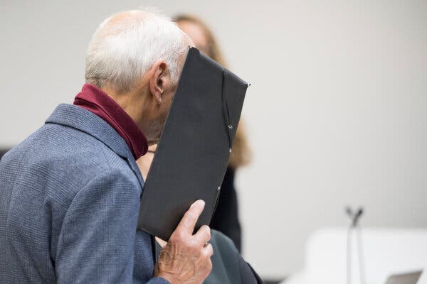 A white-haired man with a turtleneck and clue jacket holds a dark portfolio in front of his face.   