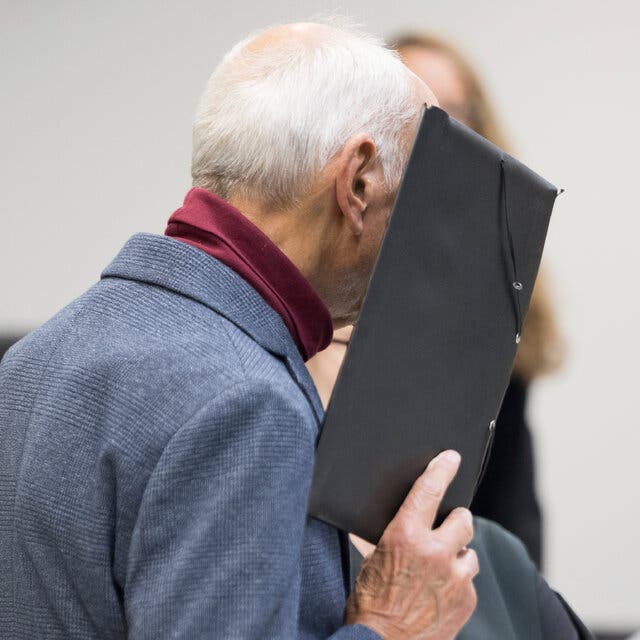 A white-haired man with a turtleneck and clue jacket holds a dark portfolio in front of his face.   