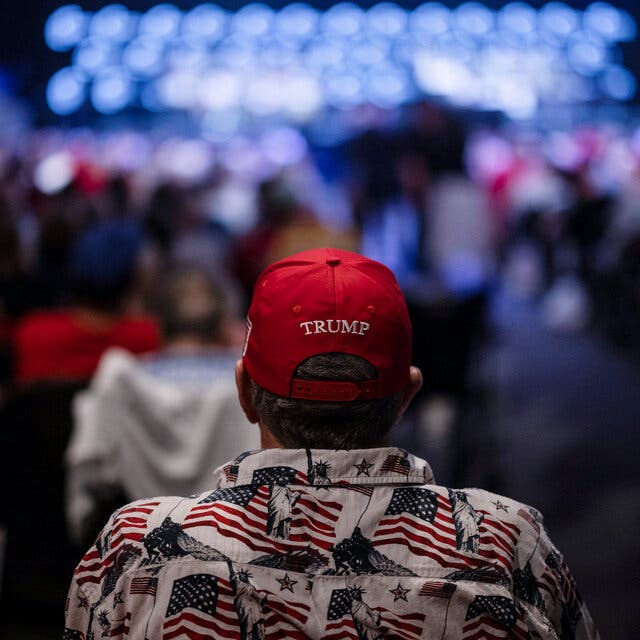 A view from behind of a man wearing a red Trump hat and a shirt decorated with American flags.