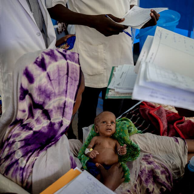 A 3-month-old baby is held by her mother while several doctors in white gowns holding notebooks surround her.