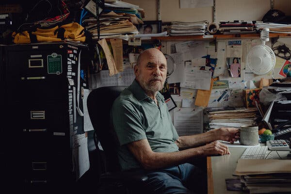 A bald man in a green polo shirt sits at a desk with a bulletin board crammed with notices behind him.